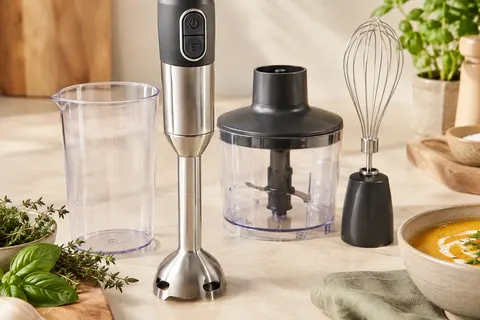 Mixeur plongeant à pied inox présenté avec son bol, son hachoir et son fouet sur un plan clair.